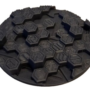 Op de afbeelding: Een zwarte, cirkelvormige basis met een getextureerd oppervlak. Het oppervlak is bedekt met een patroon van verhoogde zeshoekige vormen. De basis lijkt te zijn gemaakt van een vast materiaal, mogelijk plastic of hars.