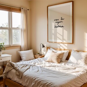 May include: A sunlit bedroom with a wooden bed frame and white bedding. A large framed print on the wall displays the text "eres tu mejor historia." A small side table holds a lamp and a plant. A window with sheer curtains is visible.
