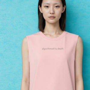 May include: A sleeveless pink top with the text "algorithmed to death" in a gray font. The top is worn by a person with short dark hair and a light blue background.