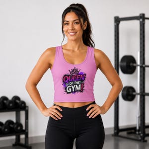 May include: A light pink sleeveless crop top featuring a graphic print with a crown icon and the text "QUEEN OF THE GYM!" in purple, black, and white lettering. The garment has a scoop neckline and a cropped hemline that sits above the waist.