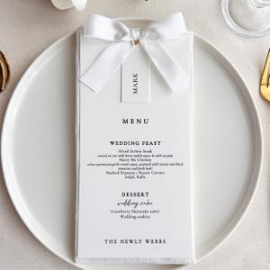 Könnte beinhalten: Eine weiße Hochzeitsmenükarte mit einer weißen Schleife und einem Etikett mit der Aufschrift "MARK". Die Speisekarte listet ein Hochzeitsmahl mit geschnittenem Sirloin-Steak, Hähnchen, Kartoffelpüree und Dessert auf. Auf der Unterseite der Karte steht "THE NEWLY WEBBS".