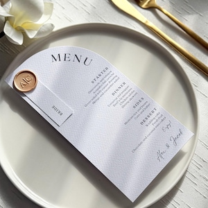 May include: A white arched menu card with black text listing starters, dinner, sides, and dessert. It features a gold wax seal with the initials AC and a small tag labeled BRIDE. The menu is printed with the names Alex & Jacob at the bottom.