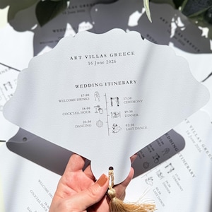May include: A white wedding itinerary card in a fan shape, featuring the text "ART VILLAS GREECE 16 June 2026" and a schedule with times for "Welcome Drinks," "Ceremony," and "Dancing." A gold tassel is attached.