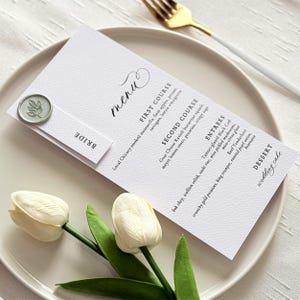 May include: A white wedding menu card with elegant script, featuring a menu with multiple courses. A small, round, green wax seal and a card that says "BRIDE" are attached. Two white tulips and a gold fork are also visible.