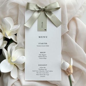 May include: A white rectangular menu card featuring black text listing courses including Sonoma Greens Salad, Filet Mignon, and Wedding Cake. A sage green satin ribbon is tied at the top with a matching tag labeled Tyler with a small cow icon.