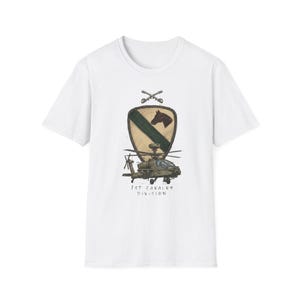 Puede incluir: Camiseta blanca con una ilustración detallada de un helicóptero militar, un escudo con una cabeza de caballo y espadas cruzadas. El texto "1st Cavalry Division" está impreso debajo del helicóptero.