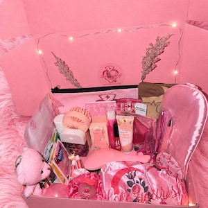 Op de afbeelding: Een roze geschenkdoos gevuld met diverse zelfzorgartikelen. De doos bevat een teddybeer, een kleine ventilator, lippenbalsem, handcrème, een slaapmasker en andere roze accessoires. De doos staat op een roze, pluizig oppervlak.
