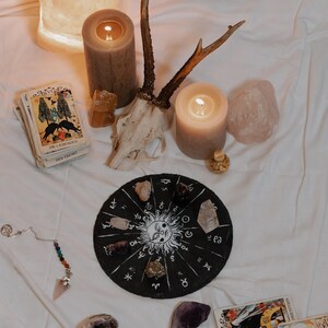 May include: A mystical arrangement featuring lit candles, crystals, tarot cards, and a decorative zodiac wheel. A deer skull and antlers are also present, along with a pendulum and a glowing salt lamp, creating a spiritual ambiance.