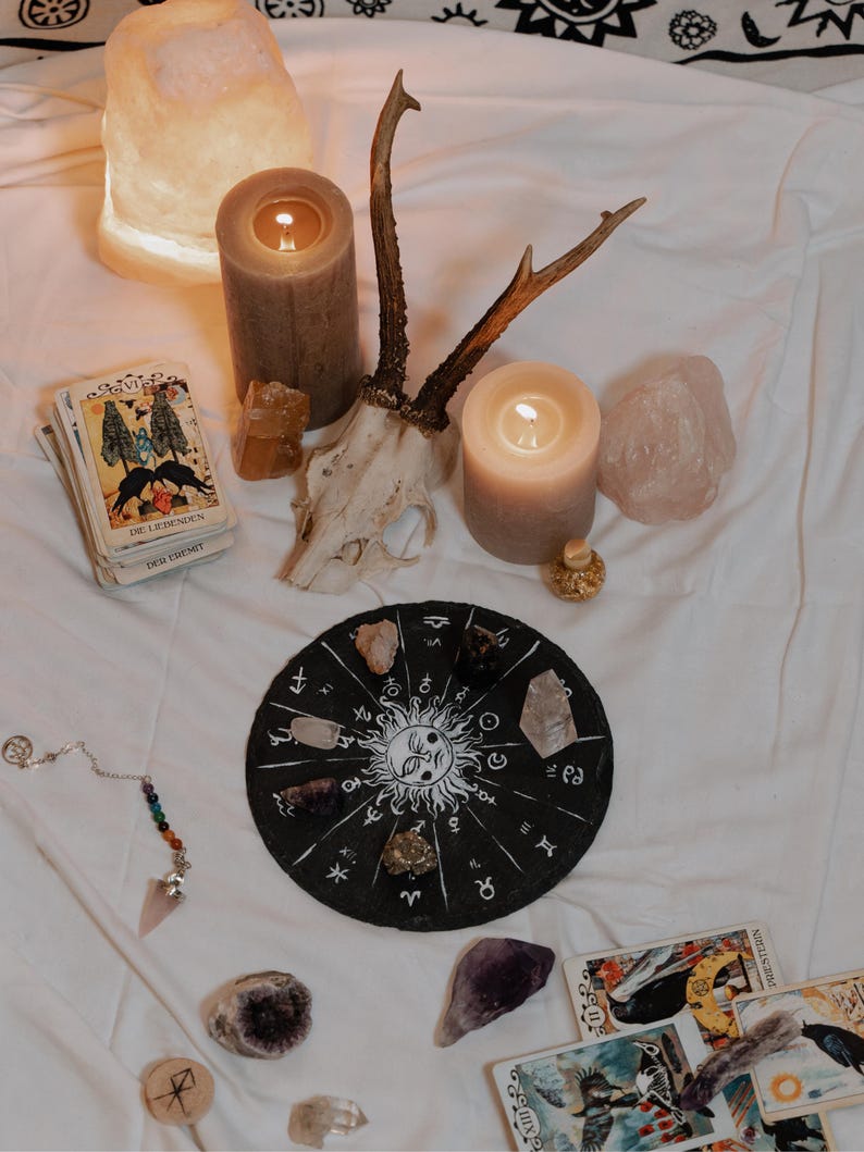 May include: A mystical arrangement featuring lit candles, crystals, tarot cards, and a decorative zodiac wheel. A deer skull and antlers are also present, along with a pendulum and a glowing salt lamp, creating a spiritual ambiance.