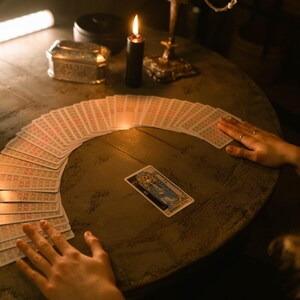 May include: A dimly lit scene featuring a tarot card reading. A deck of tarot cards is fanned out on a round wooden table. A lit black candle and a gold candelabra are also on the table. The hands of a person are near the cards.
