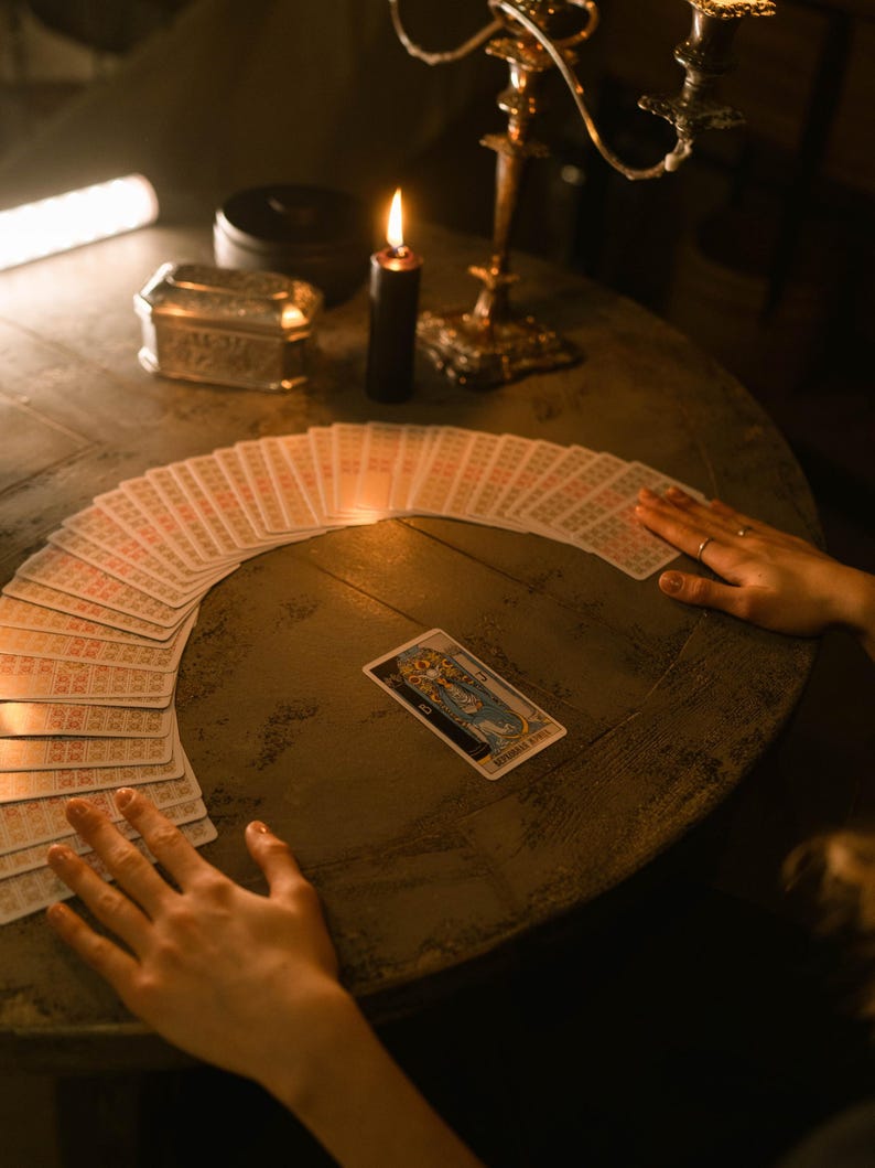 May include: A dimly lit scene featuring a tarot card reading. A deck of tarot cards is fanned out on a round wooden table. A lit black candle and a gold candelabra are also on the table. The hands of a person are near the cards.