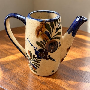 May include: A cream-colored ceramic pitcher featuring a hand-painted floral design in shades of dark blue, green, and brown. The pitcher has a curved handle, a narrow spout, and a blue rim. The surface has a speckled texture throughout the body.