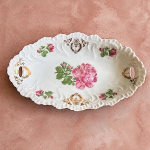 Puede incluir: Una bandeja de servicio ovalada de porcelana blanca con bordes festoneados y dos asas integradas. El centro presenta una gran rosa rosa con hojas verdes, rodeada de motivos florales y detalles de filigrana dorada.