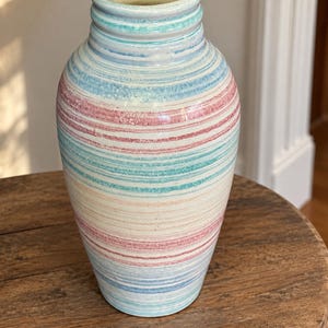 May include: A ceramic vase with a cylindrical body and a narrow neck, featuring a horizontal striped pattern in shades of light blue, soft pink, and turquoise against a cream-colored background. The surface has a smooth, glazed finish.