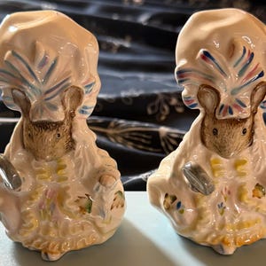 May include: Two ceramic figurines depicting anthropomorphic mice dressed in elaborate period clothing with ruffled bonnets. The mice are painted in cream, brown, blue, and pink tones, featuring detailed textures on their garments and hats.