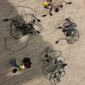 May include: Four separate sets of string lights with dark green or tan wires and black sockets. Each set is tangled in a loose pile. One set features small red beads along the wire, while another is bundled with tan tape and a black electrical plug.