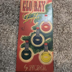 Puede incluir: Caja de luces navideñas vintage con el texto "GLO-RAY Lights by NOMA". La caja presenta un fondo rojo e ilustraciones de luces navideñas en rojo, azul, verde y amarillo. La caja rectangular tiene un diseño retro.