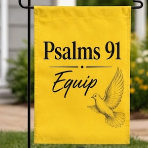 May include: Yellow garden flag with the text "Psalms 91 Equip" in black script. A yellow dove illustration is featured below the text. The flag is hanging on a black metal stand, with a blurred green background.