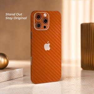 Puede incluir: Funda de teléfono naranja con textura de fibra de carbono con el logotipo de Apple. La funda tiene un módulo de cámara con múltiples lentes. El texto "Stand Out Stay Original" es visible en el lado izquierdo de la imagen.