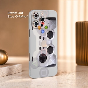 May include: A clear smartphone case featuring a graphic print of a video game controller with colorful buttons and analog sticks. The text Stand Out Stay Original is printed in white on the upper left side. The case has a cutout for the camera lens.