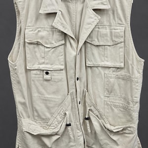 May include: A beige utility waistcoat featuring a collared neckline, button-down front, and multiple cargo-style pockets on the chest and waist. The garment includes shoulder epaulets and a label reading "Tiky Coton DOCTOR 100% COTON".