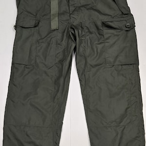 May include: Dark olive green cargo trousers featuring a wide-leg silhouette, a fabric waist tie, and two large side pockets with buttoned flaps. The trousers include reinforced knee panels and adjustable buttoned cuffs at the bottom of each leg opening.