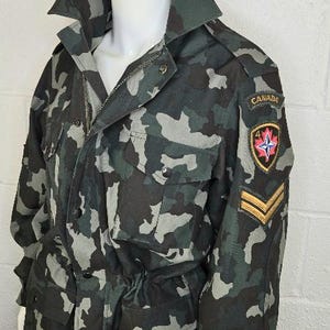 Peut inclure: Veste de camouflage avec un col, plusieurs poches et une taille ajustable. La veste présente un écusson "CANADA" avec une feuille d'érable et un emblème d'étoile, ainsi que des rayures dorées sur la manche. Le mannequin porte également des lunettes de soleil à monture dorée.