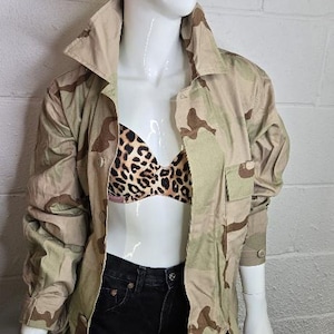 Peut inclure: Veste de camouflage beige et marron avec deux poches avant et une fermeture boutonnée. La veste est associée à un soutien-gorge à imprimé léopard et à un jean noir. Le mannequin est présenté devant un mur de briques blanches.