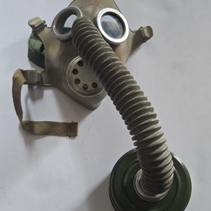 Peut inclure: Un masque à gaz en caoutchouc beige doté de deux oculaires circulaires en verre avec des bords en métal argenté, une valve centrale circulaire et un long tuyau gris flexible et nervuré relié à un filtre cylindrique en métal vert foncé.