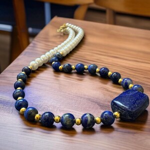 May include: A necklace featuring a combination of white pearls, deep blue lapis lazuli beads, and gold-colored accents. The necklace includes a large, rectangular lapis lazuli stone pendant. The jewelry is displayed on a wooden surface.