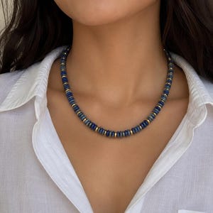 May include: A beaded necklace featuring alternating small, round, deep blue lapis lazuli stones and thin, polished gold-tone metal spacers. The necklace sits close to the base of the neck, showcasing a repeating pattern of blue beads separated by single gold discs.