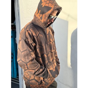 Puede incluir: Una sudadera con capucha de manga larga con un patrón de panal en negro y naranja quemado. La prenda incluye una capucha con cordón, un bolsillo canguro delantero y puños acanalados. El diseño general muestra un motivo hexagonal geométrico.