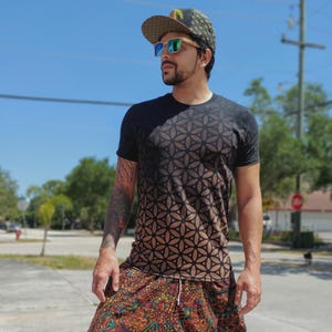 May include: A short-sleeved black t-shirt featuring a repeating geometric flower of life pattern that fades into a brown hue at the bottom. The outfit includes a baseball cap with a matching geometric print and sunglasses with teal lenses.