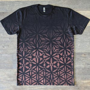 May include: A black short-sleeve t-shirt featuring a repeating geometric flower of life pattern that transitions from a dark black at the top to a faded, muted reddish-brown hue toward the bottom hem, with a standard crew neckline and a neck label.