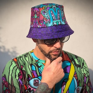 May include: A bucket hat featuring a vibrant, multi-coloured abstract pattern in shades of purple, teal, and magenta, with a dark purple brim adorned with a repeating geometric hexagon print, paired with a matching short-sleeved graphic shirt.