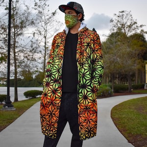 May include: A long-sleeved, knee-length coat featuring a vibrant, repeating geometric star pattern in shades of orange, yellow, and green with black outlines. The coat is worn over a plain black shirt and dark trousers, paired with a matching patterned face mask.