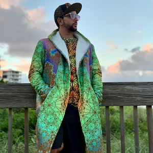 May include: A long hooded coat featuring a vibrant green, yellow, and blue geometric fractal pattern with a white interior lining. The coat is worn over a short-sleeved shirt with a black and orange geometric print, dark shorts, and a patterned cap.