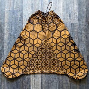 Op de afbeelding: Mosterdgele en donkerbruine harembroek met een geometrisch honingraatpatroon aan de zijkanten en een cirkelvormig bloemenmandala-ontwerp op het middenpaneel. De broek heeft een elastische tailleband met trekkoord en twee zijzakken.