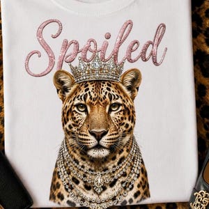 Op de afbeelding: Een wit t-shirt met een afbeelding van een luipaard die een met juwelen bezette kroon en meerdere gelaagde kristallen kettingen draagt. Boven de luipaard staat het woord "Spoiled" gedrukt in een roze glitter-schrijflettertype.
