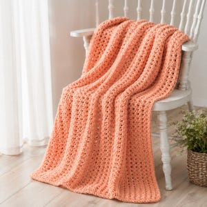 May include: A peach-colored crochet throw blanket featuring a repeating vertical ribbed pattern with openwork stitches. The textile is draped over a white wooden chair, showcasing a soft, textured yarn construction with consistent, uniform loops.