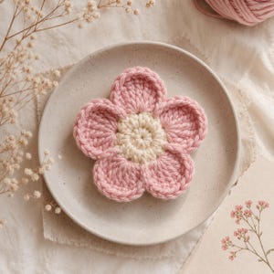 Pode incluir: Uma flor de crochê feita à mão com cinco pétalas rosas arredondadas que envolvem um núcleo circular de cor creme. A peça têxtil tem formato circular e apresenta uma textura de fio consistente em todo o design floral.