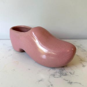 May include: A pink ceramic clog-shaped planter. The planter has a rounded toe and heel, with a hollowed-out interior for plants. The surface has a speckled texture and a glossy finish. The clog is a traditional Dutch design.
