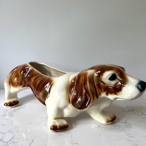 May include: A ceramic dog planter in a brown and white color scheme. The dog is in a standing position with a long body and a large opening on its back for plants. The dog has black eyes and a glossy finish.