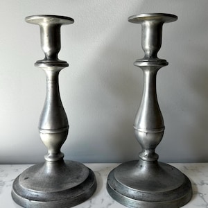 May include: Two antique silver-colored candlesticks. Each candlestick has a round base, a slender, curved stem, and a circular top designed to hold a candle. The candlesticks are displayed on a white marble surface.