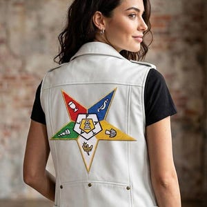 Pode incluir: Um colete de couro branco com um grande emblema de estrela colorida nas costas. O emblema apresenta vários símbolos em vermelho, azul, amarelo, verde e branco. O colete tem um estilo motociclista clássico com gola e costuras visíveis.