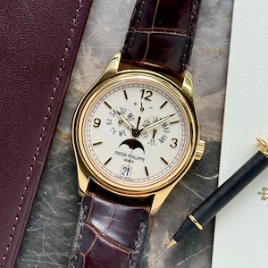 May include: A gold-toned wristwatch with a white face, featuring black hands and hour markers. The watch has a brown leather band with a crocodile pattern. The watch face displays the date and day of the week. A black pen rests beside the watch.
