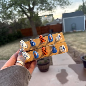 May include: A wooden display board holding several sets of painted artificial nails. The nails feature designs with the letters "OKC", lightning bolts, basketballs, and the numbers "2" and "3". The nails are painted in blue, orange, white, and yellow.