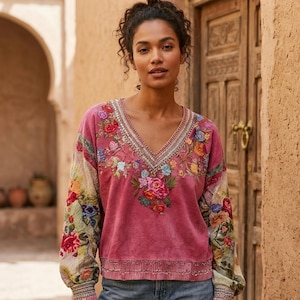 Puede incluir: Una blusa rosa con cuello en V y bordado floral. Las mangas tienen un estampado floral colorido. El escote y los puños están adornados con costuras decorativas. La blusa está hecha de una tela suave y texturizada.