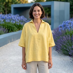 Op de afbeelding: Een vrouw draagt een gele linnen top met een V-hals en korte mouwen. De top wordt gecombineerd met een lichtgekleurde broek. De achtergrond toont een tuin met blauwe bloemen en een moderne architectonische structuur.