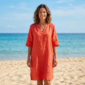May include: A knee-length, coral-colored dress with a V-neck and short sleeves. The dress is made of a lightweight fabric and is worn on a sandy beach with the ocean in the background. The dress has a relaxed fit.